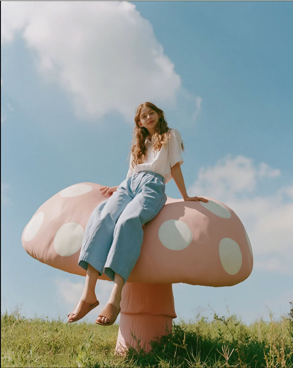 A woman with long wavy hair sitting atop a large pink mushroom with white spots in a grassy field under a blue sky.