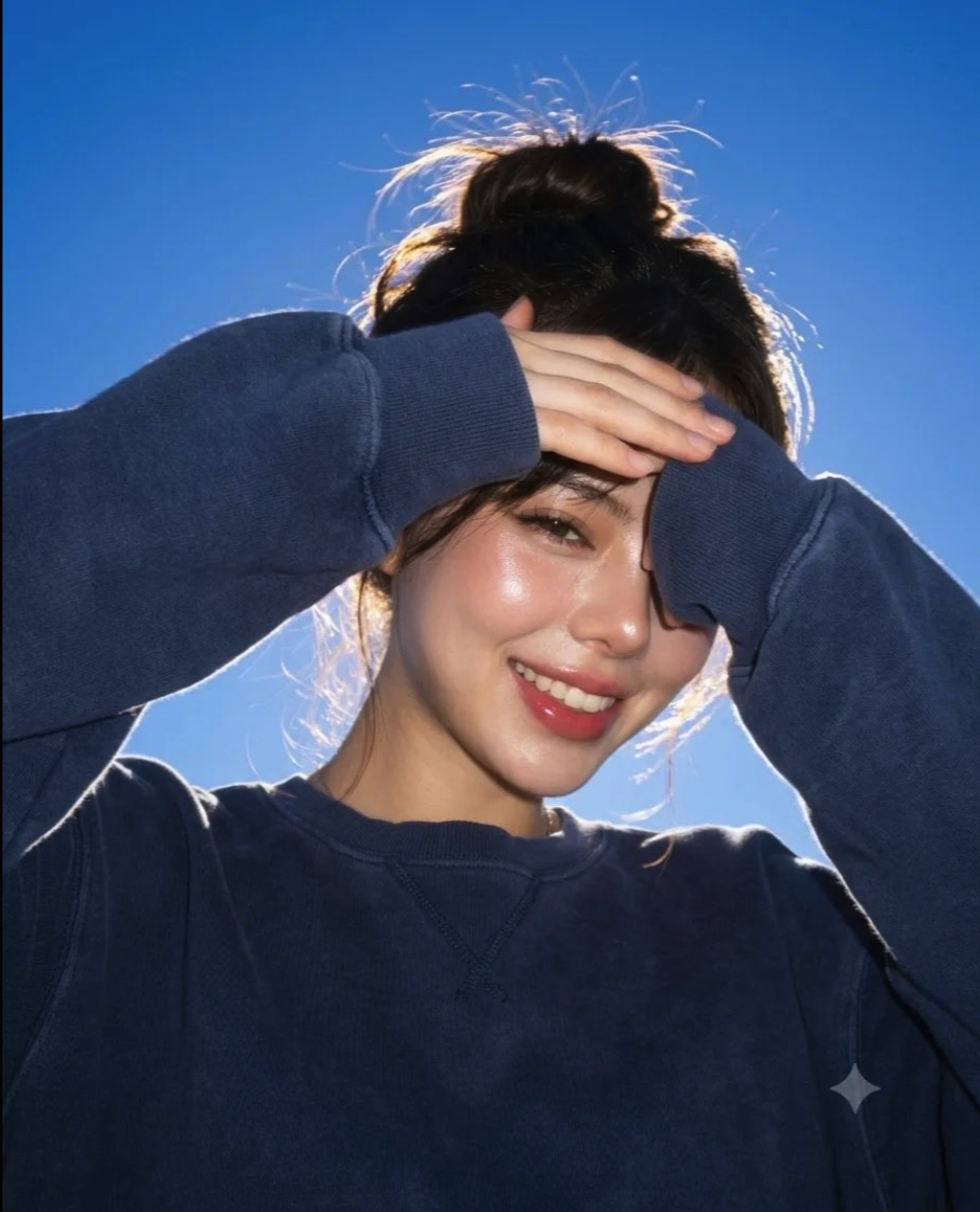 A smiling woman with dark hair in a loose bun raising her hands near her face, photographed from a low angle against a clear blue sky.