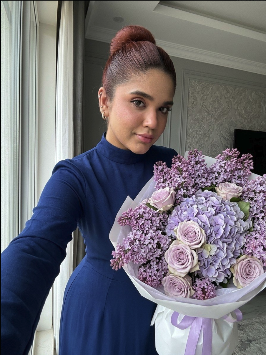 A woman with red hair in a bun, wearing a navy turtleneck dress, holding a bouquet of purple flowers.