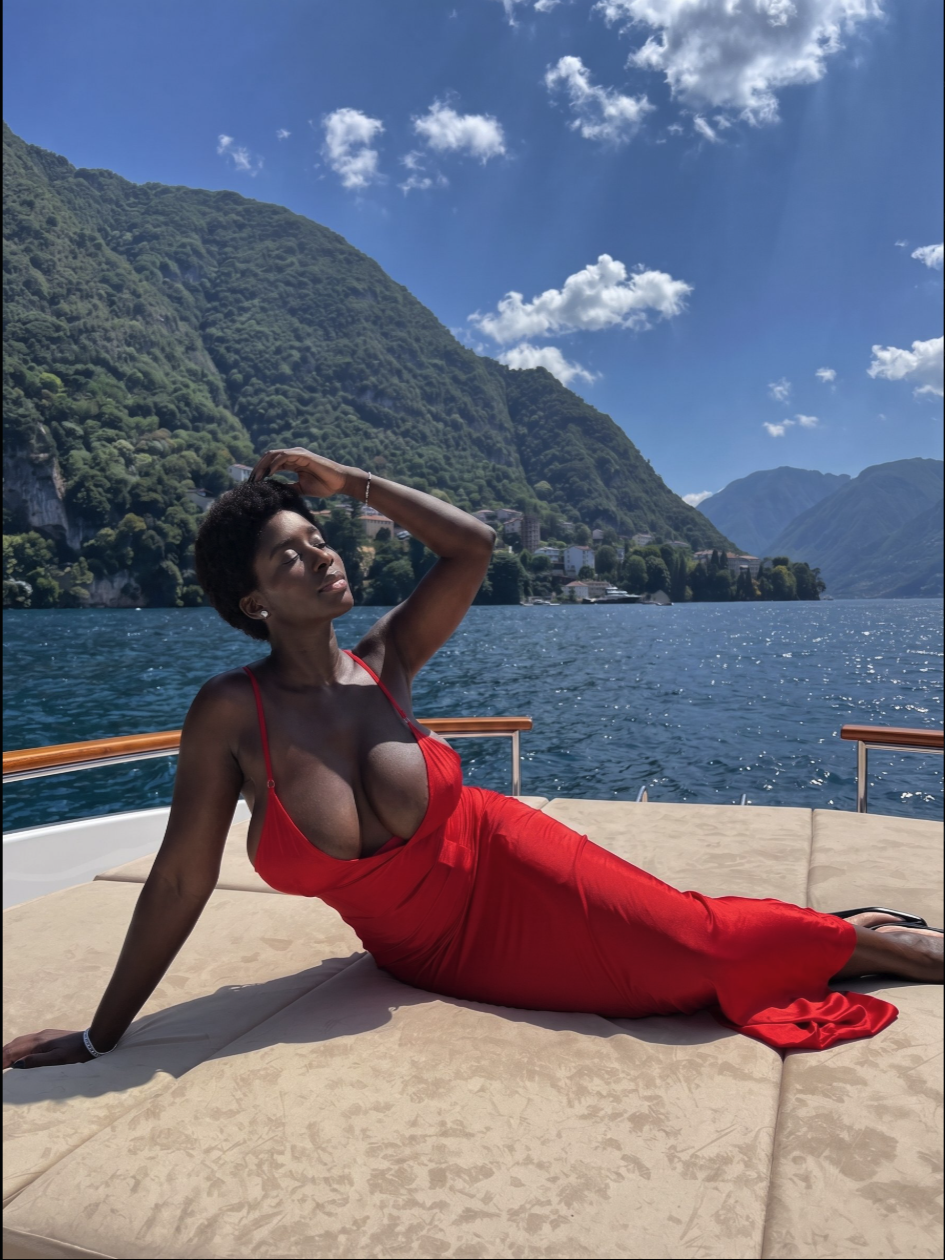 A woman in a low-cut red dress reclining on a boat deck, with forested mountains and a lake behind her.