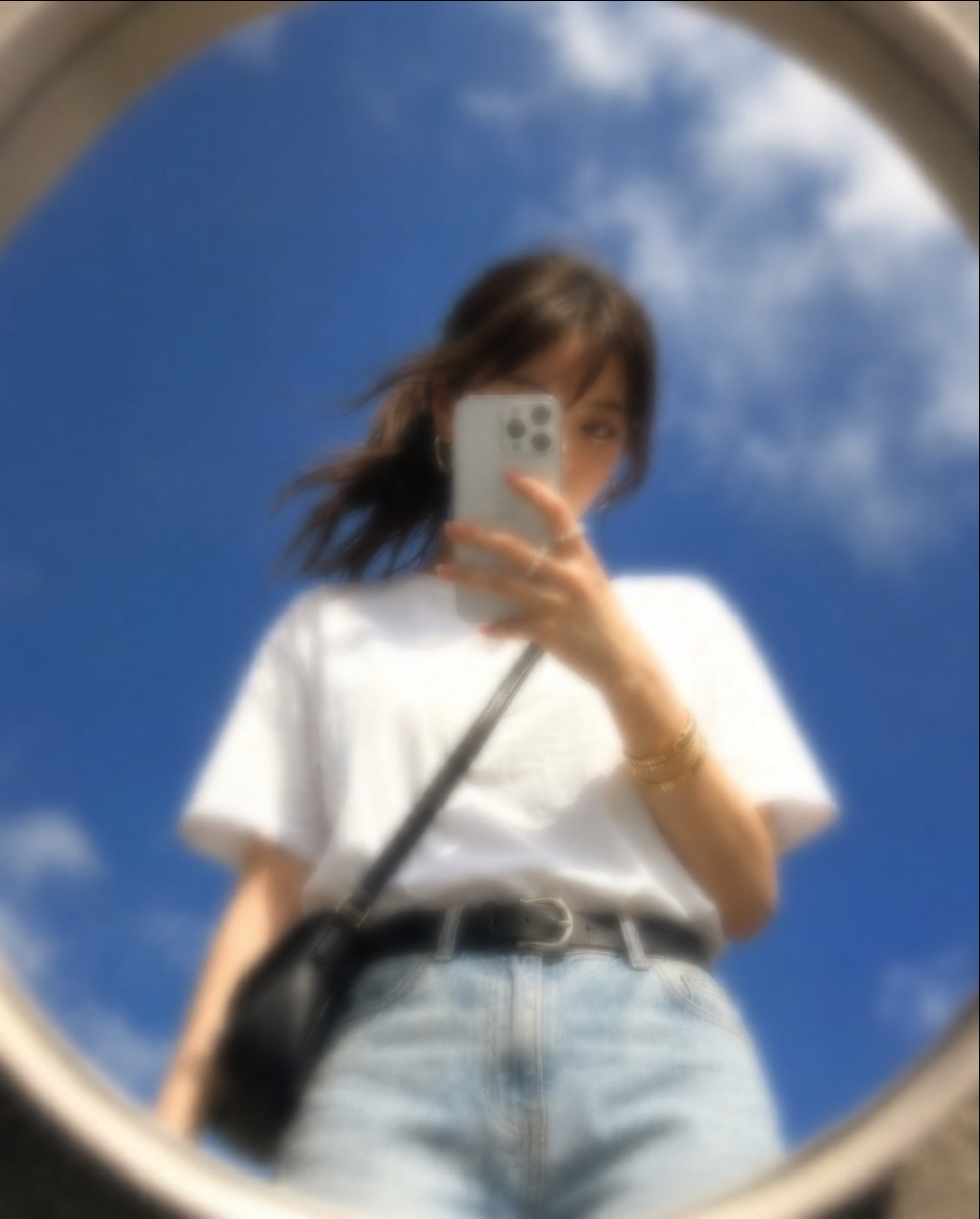A young woman taking a selfie with a white iPhone, reflected in a round mirror outdoors.