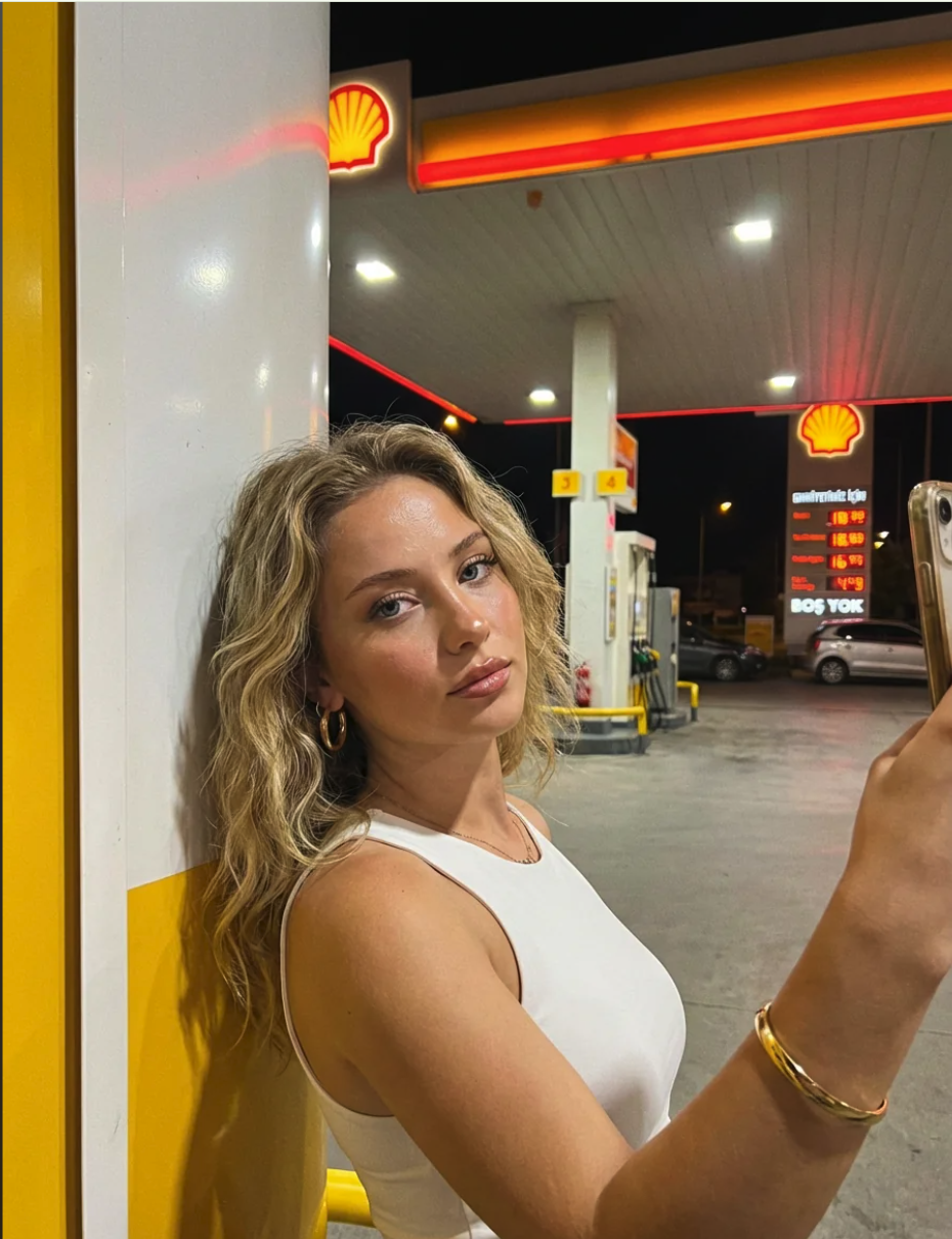 A woman with blonde wavy hair taking a selfie at a Shell gas station at night.