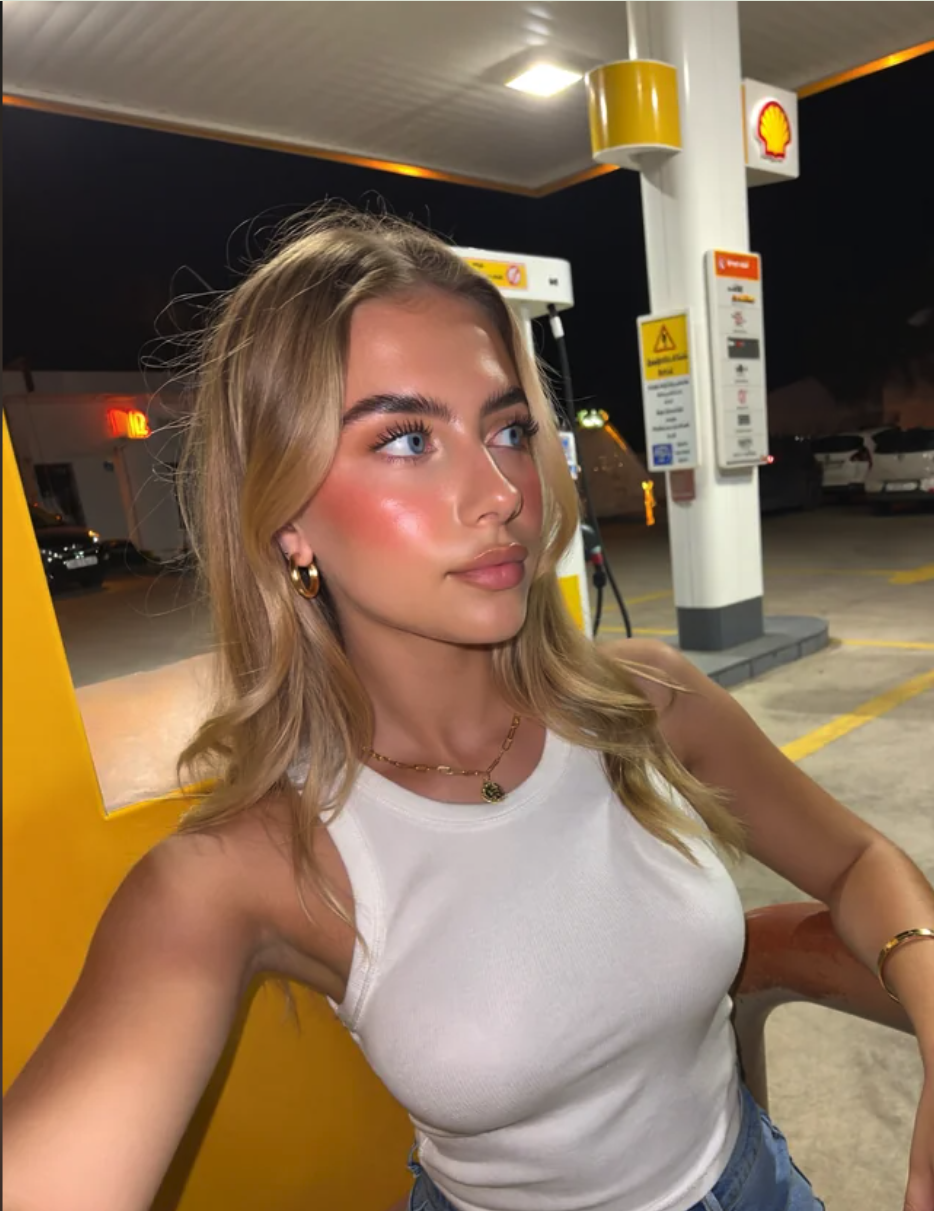 A young woman with long blonde hair at a Shell gas station at night, wearing a white tank top and gold jewelry.