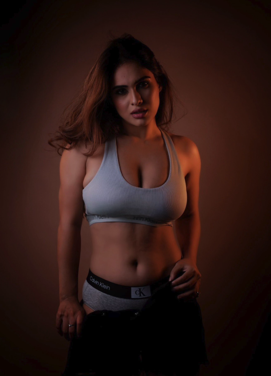A woman with dark hair wearing Calvin Klein branded sports bra and underwear, photographed in a dimly lit studio with reddish lighting.