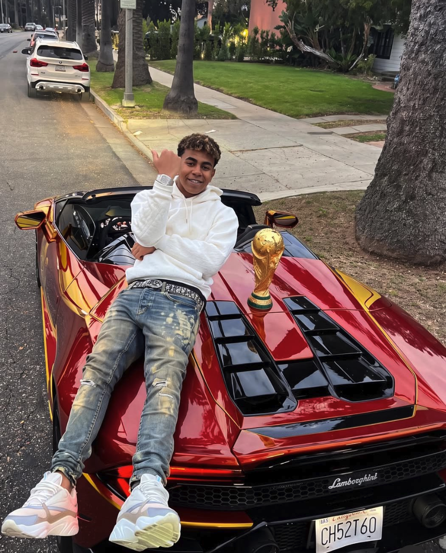 Lamine Yamal sitting on the hood of a red Lamborghini convertible beside a World Cup trophy on a residential street.