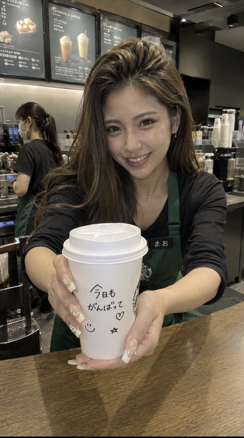 A woman wearing a green Starbucks apron and holding out a white coffee cup with handwritten Japanese writing.