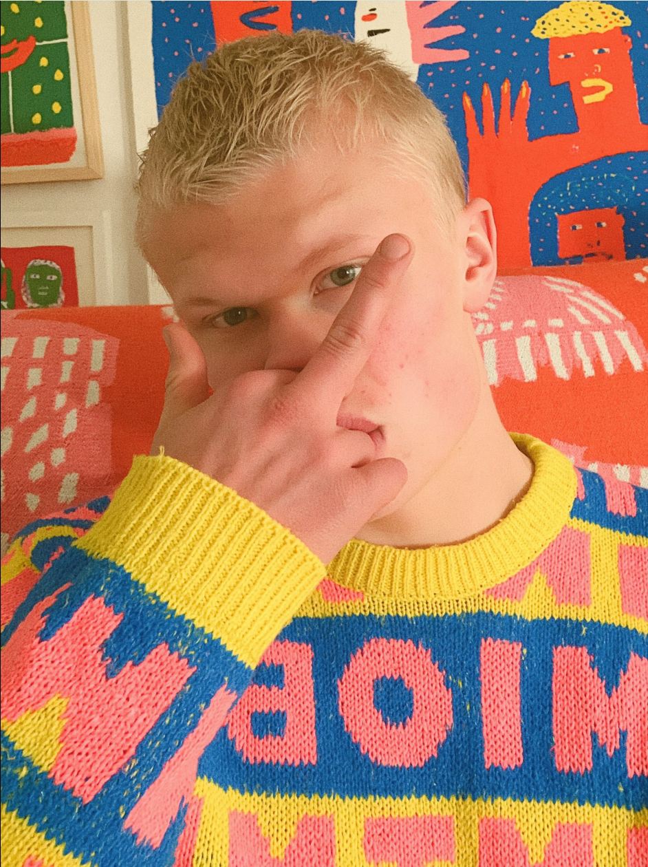 A young person with short blonde hair in a yellow, pink, and blue patterned sweater making a hand gesture over their face.