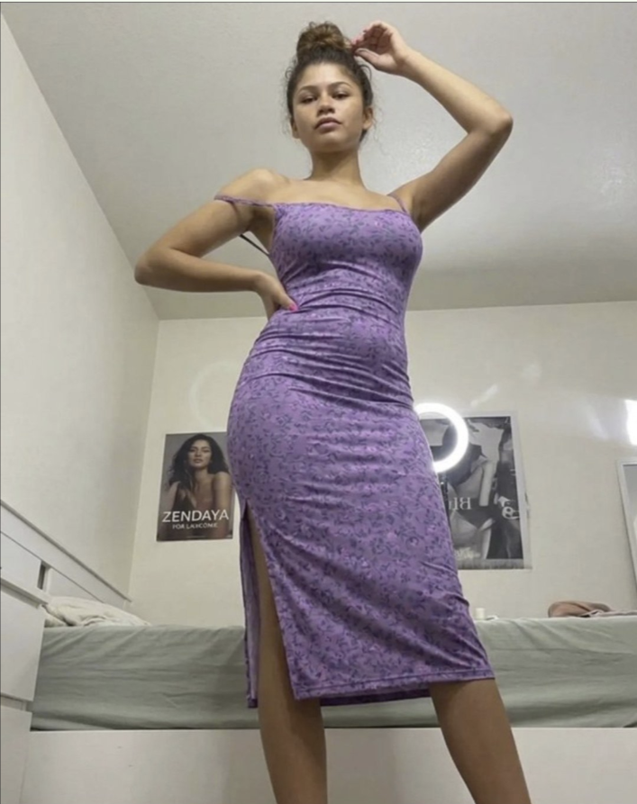 A woman resembling Zendaya posing in a fitted purple floral midi dress beside a bed in a bedroom.