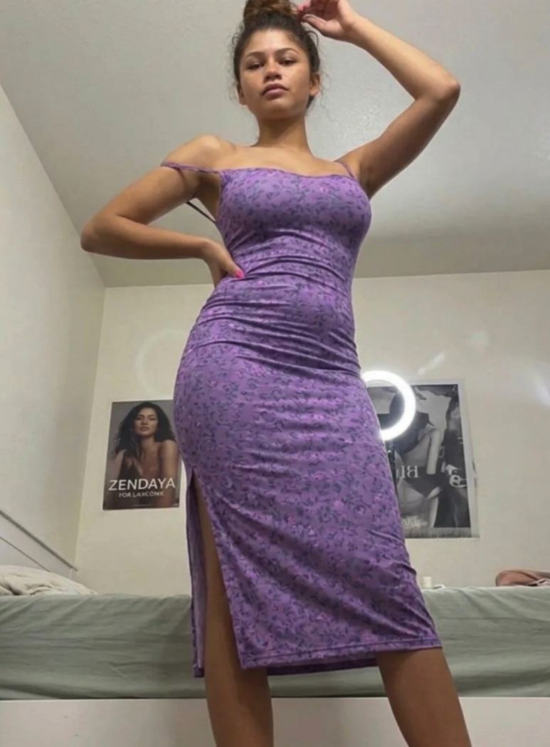 A woman resembling Zendaya standing in a bedroom wearing a fitted purple patterned dress.
