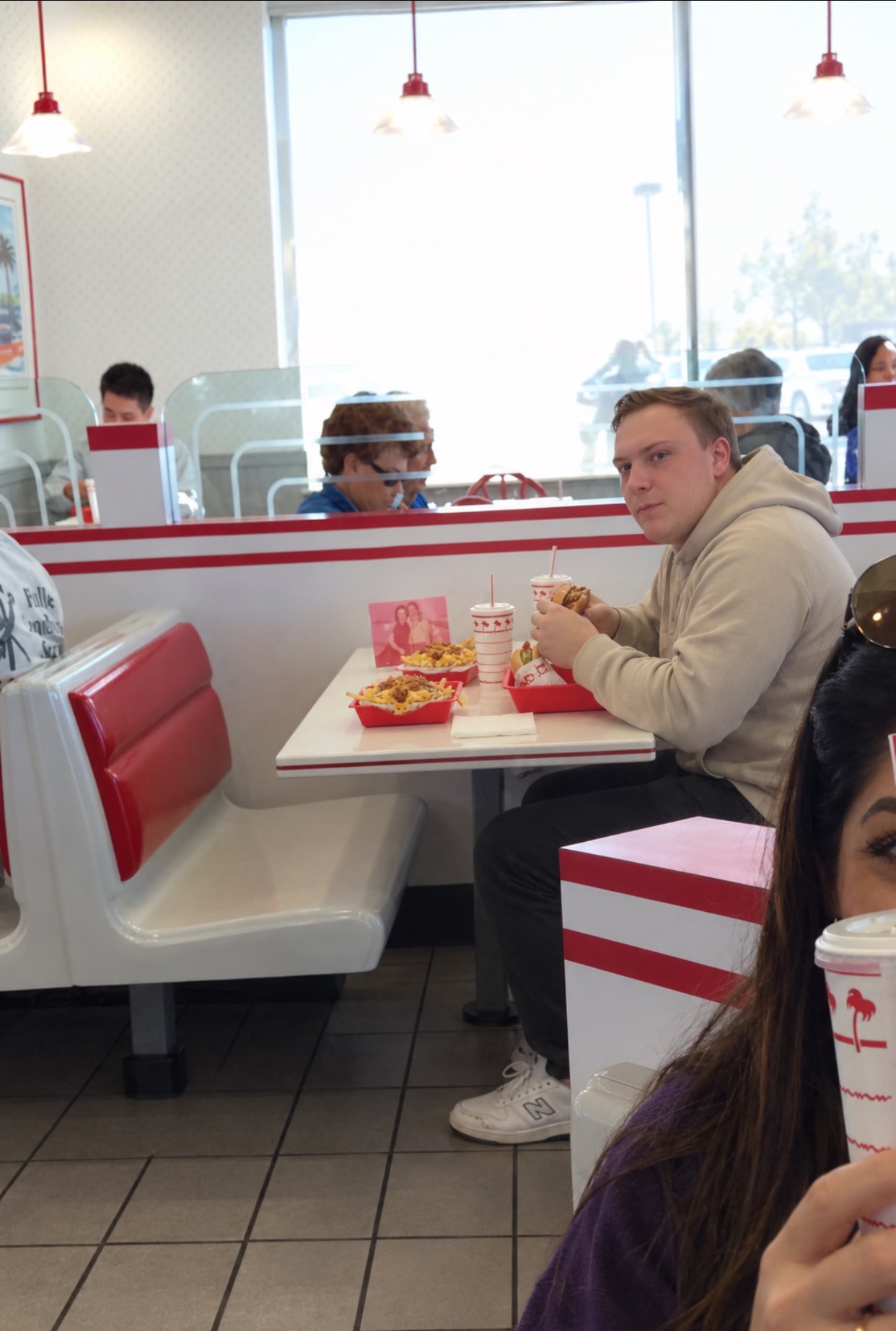 A man resembling Harrison Mevis seated in a red-and-white booth at an In-N-Out Burger, holding a burger beside fries and a drink.