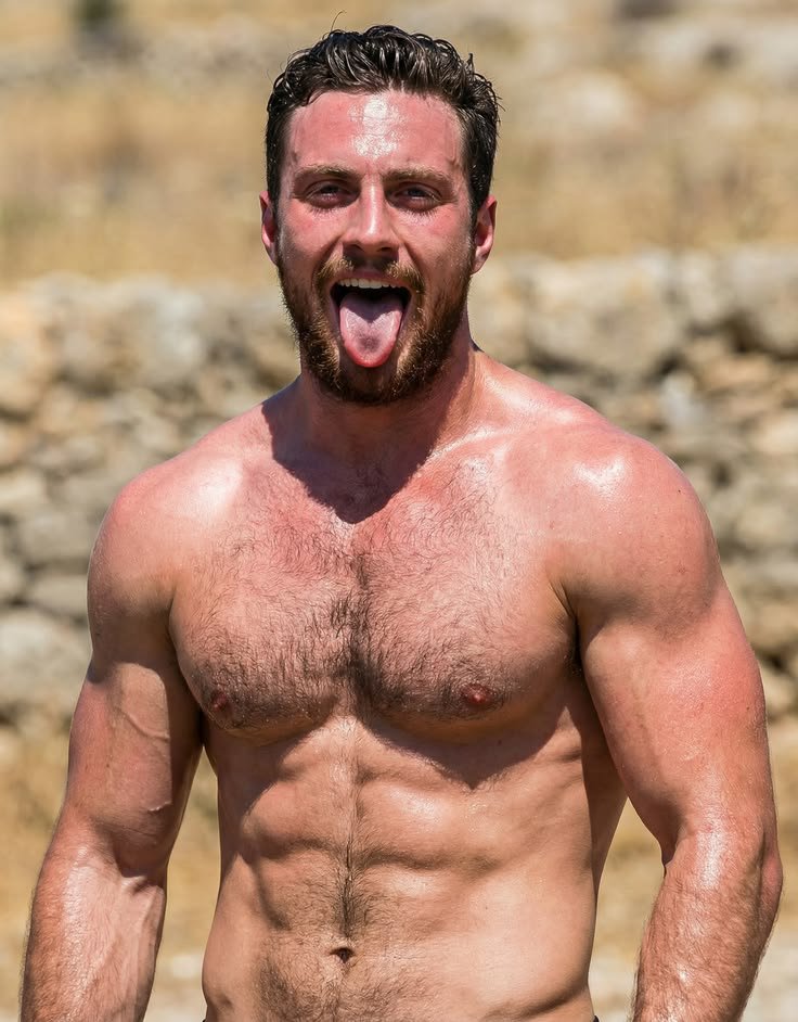 A man resembling Aaron Taylor-Johnson standing shirtless outdoors in front of a dry stone wall.