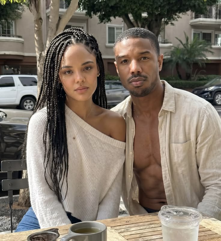 A man resembling Michael B. Jordan seated at an outdoor café table beside an unidentified woman with long braids.