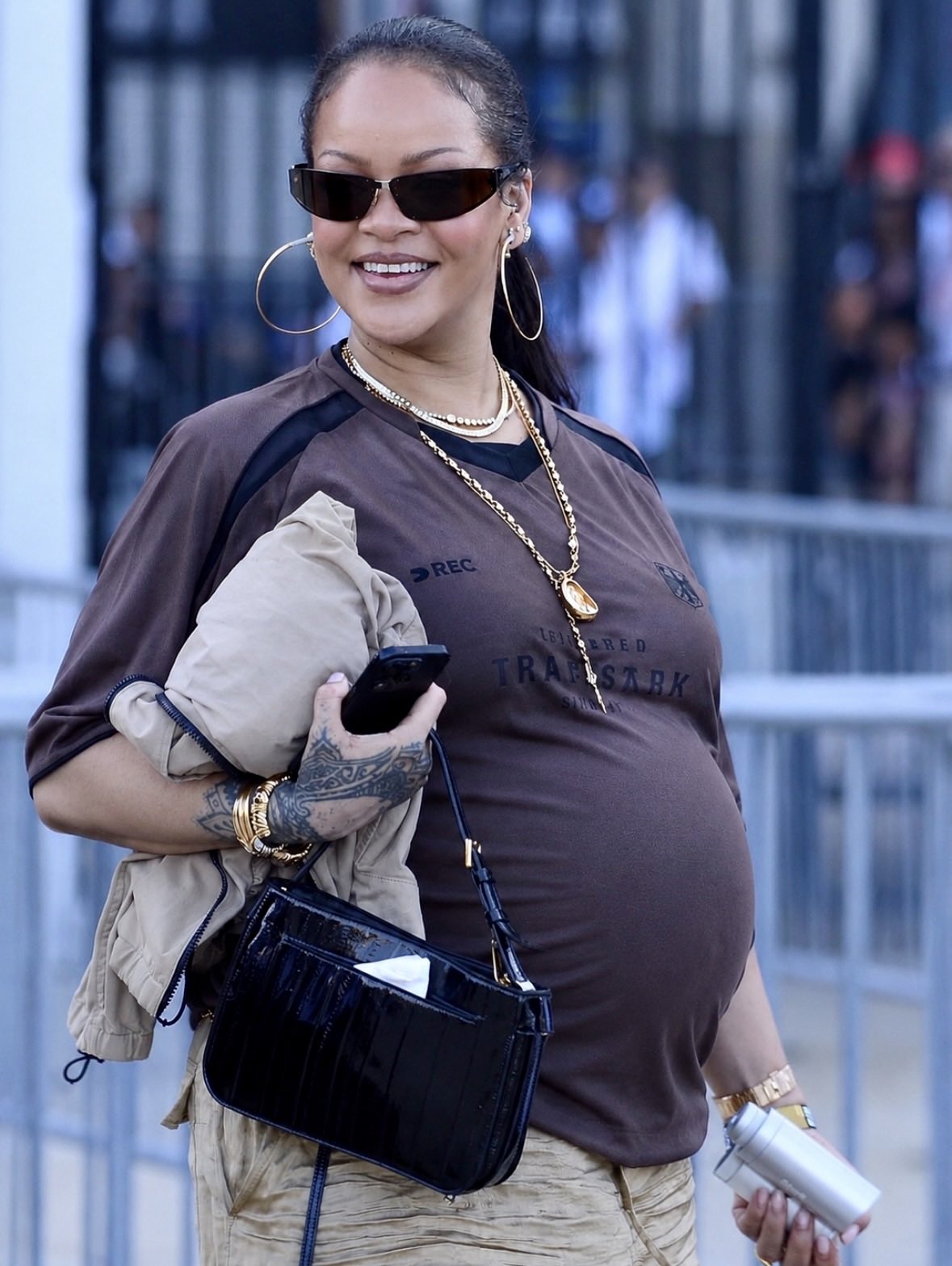 A woman resembling Rihanna, visibly pregnant and smiling, standing behind a metal barrier on a city street.