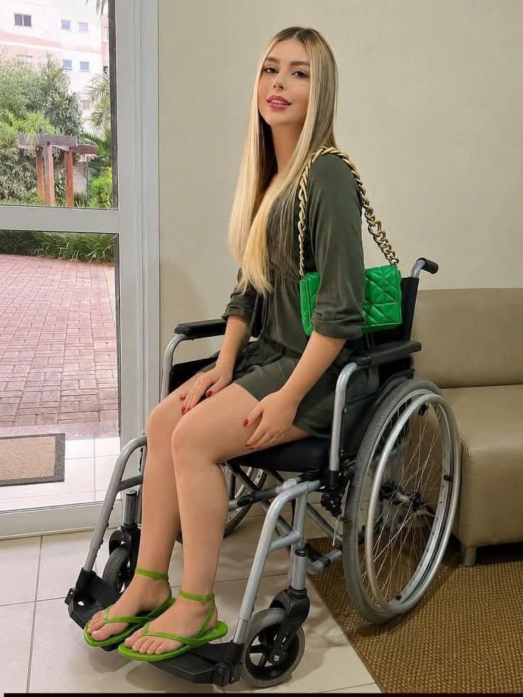 A woman with long blonde hair, wearing an olive dress, seated in a wheelchair and holding a green quilted handbag with a gold-tone chain.