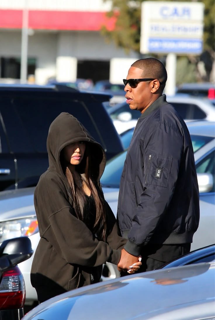 A man resembling Jay-Z standing beside a woman resembling Nicki Minaj in a hooded jacket, between parked cars at an outdoor lot.