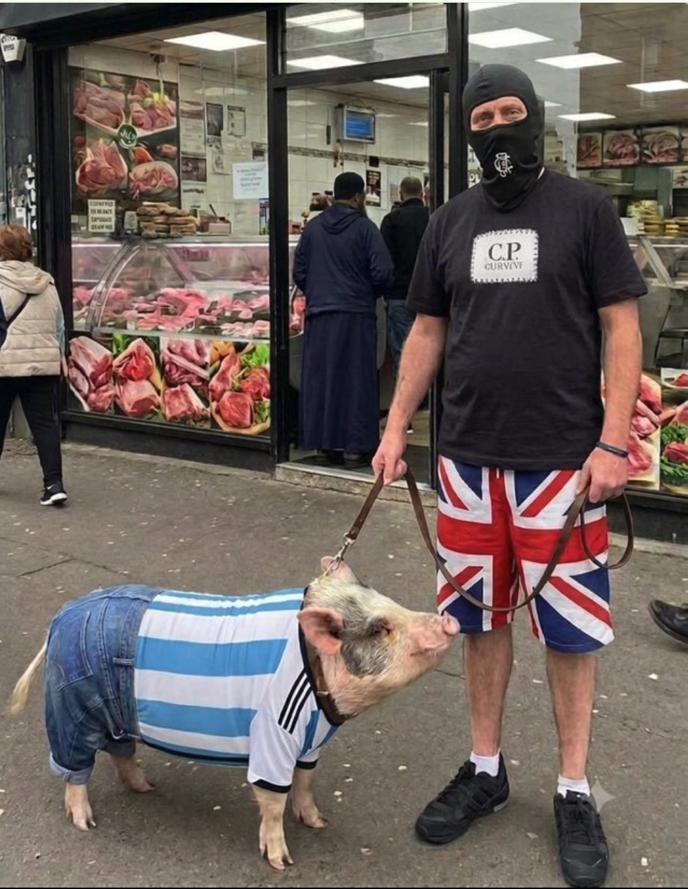 A masked man in Union Jack shorts standing with a pig wearing a striped jersey and denim shorts outside a butcher shop.