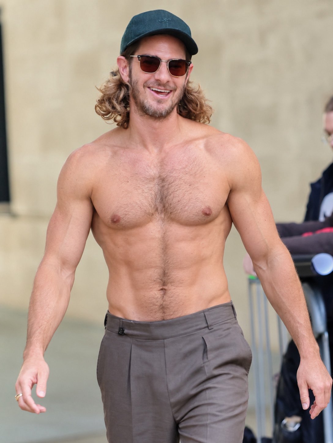 A man resembling Andrew Garfield walking shirtless in trousers, a green cap, and sunglasses.
