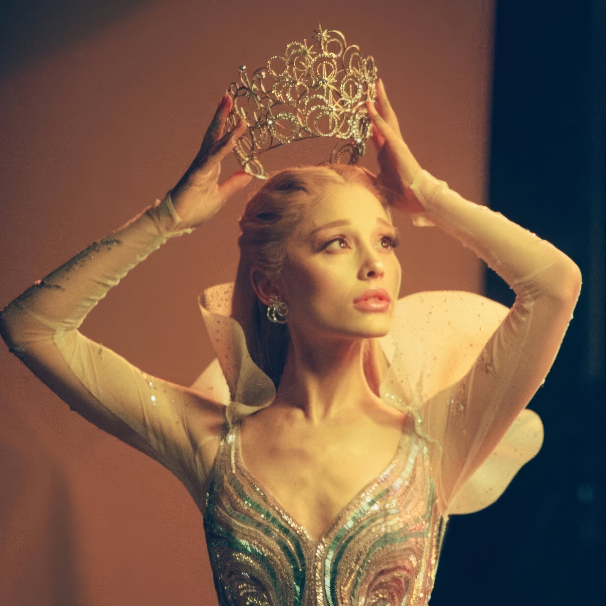 A woman resembling Ariana Grande holding a jeweled tiara above her head while wearing a beaded green and silver corset with sheer ruffled sleeves.