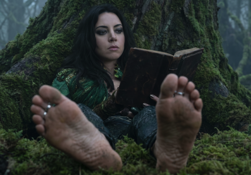 A woman resembling Aubrey Plaza in a green dress reading an old book while seated against a mossy tree, with bare feet in the foreground.