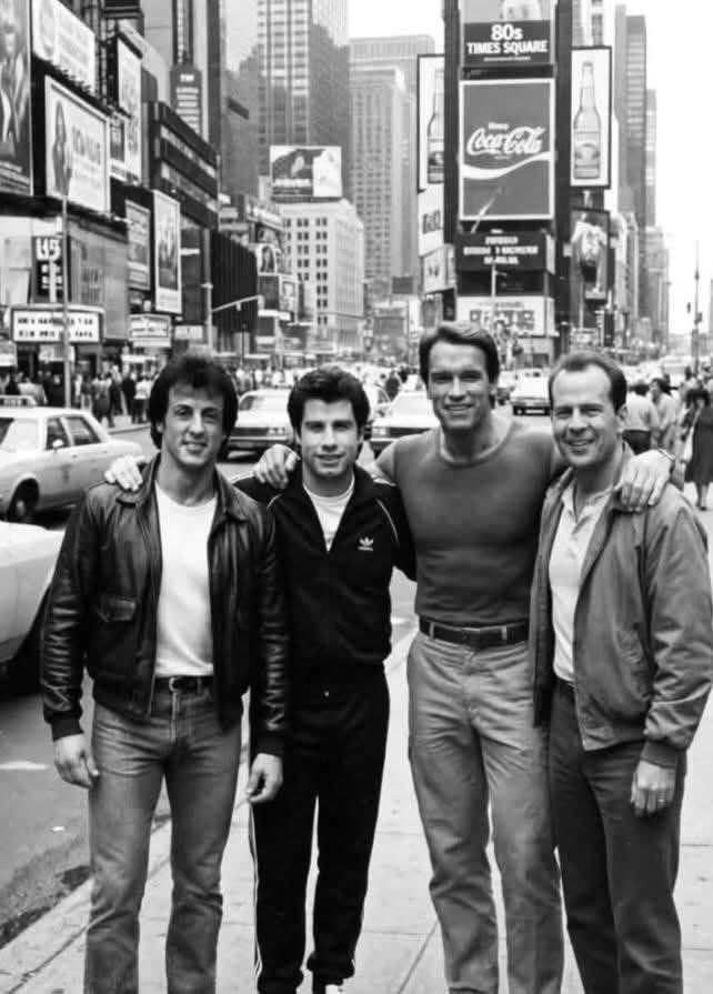 Four men resembling Bruce Willis, Arnold Schwarzenegger, Sylvester Stallone, and John Travolta on a Times Square sidewalk.