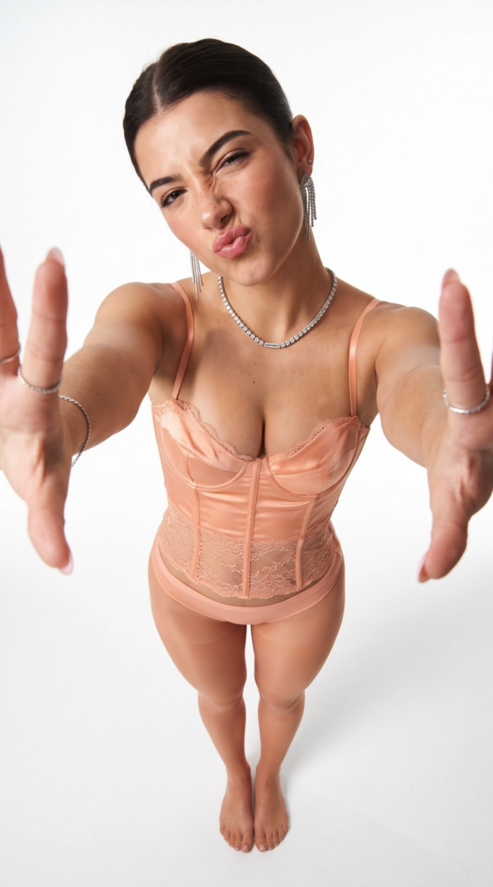 A woman resembling Charli D'Amelio posing in a peach lace corset bodysuit with her arms extended toward the camera.