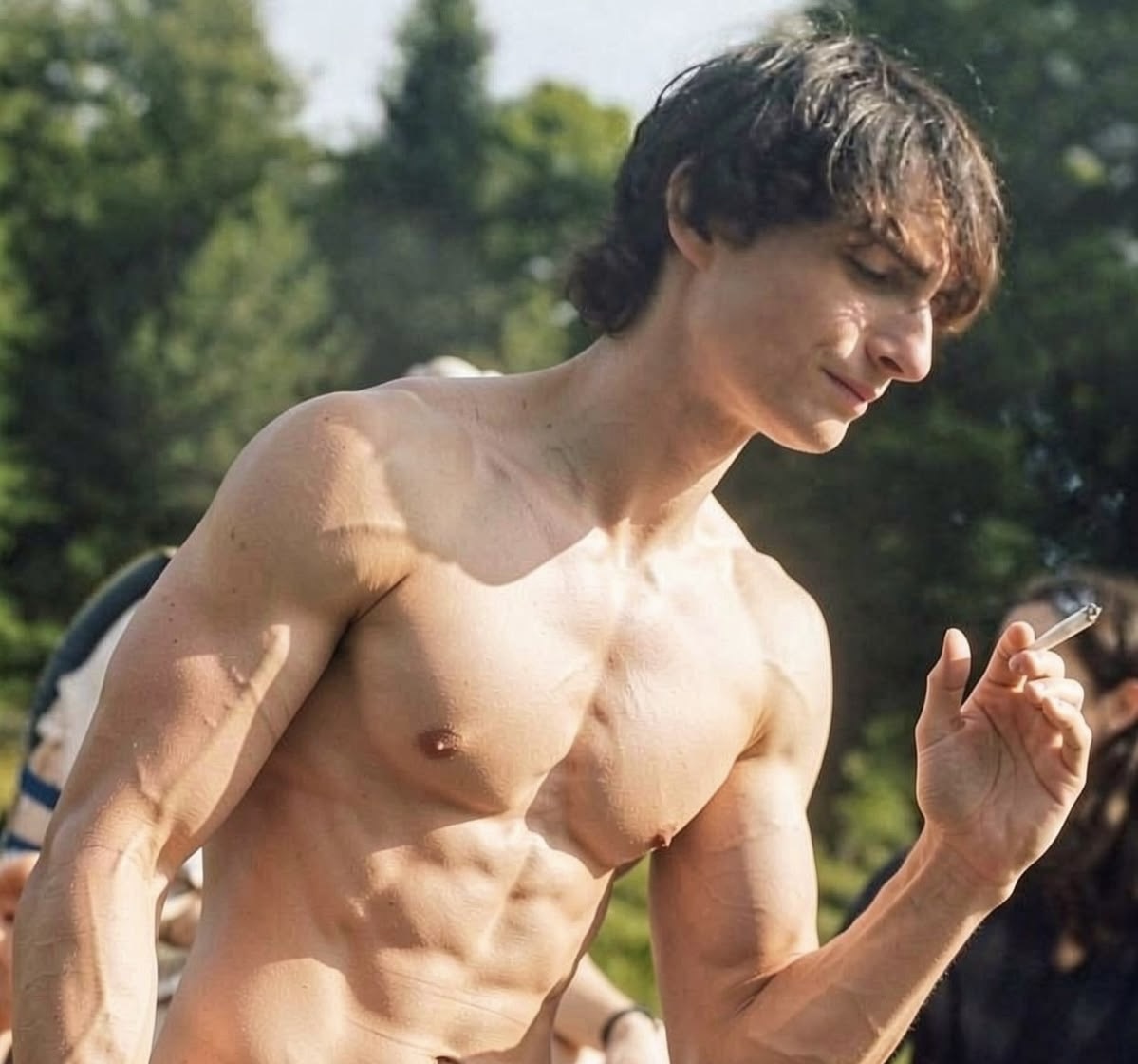 A sdeepfake of Finn Wolfhard standing outdoors among trees, looking down at a small object in his hand.