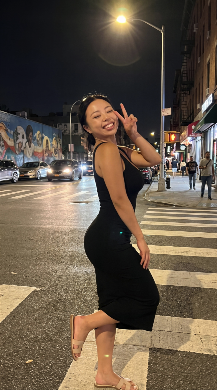 A woman in a black dress posing with a peace sign on a city crosswalk at night.