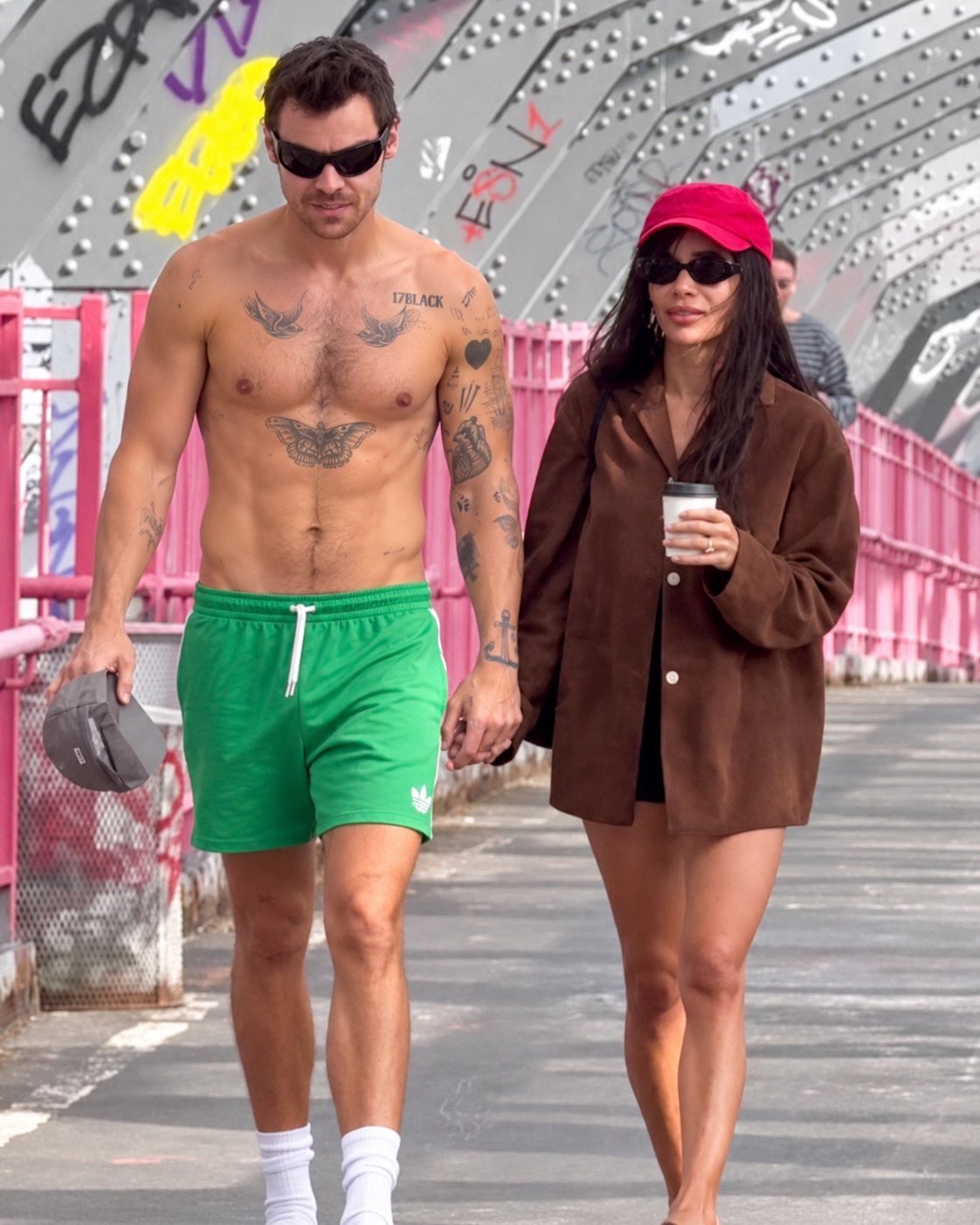 A shirtless man resembling Harry Styles and a woman resembling Zoe Kravitz walking hand-in-hand beside a graffitied metal bridge.