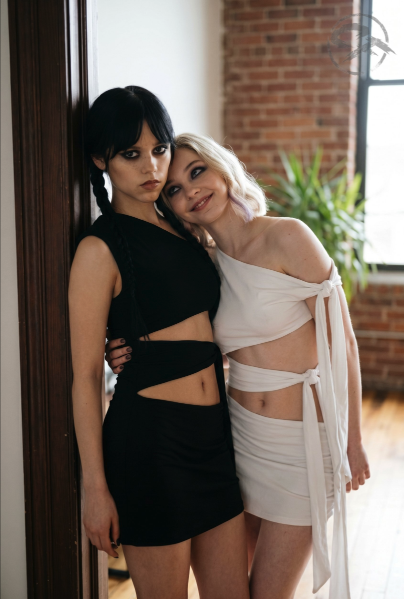 Two women resembling Jenna Ortega and Emma Myers posing together indoors against a brick wall.