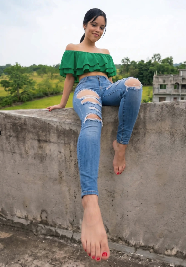 A woman resembling Jenna Ortega sitting barefoot on a concrete rooftop wall in a green crop top and ripped jeans.