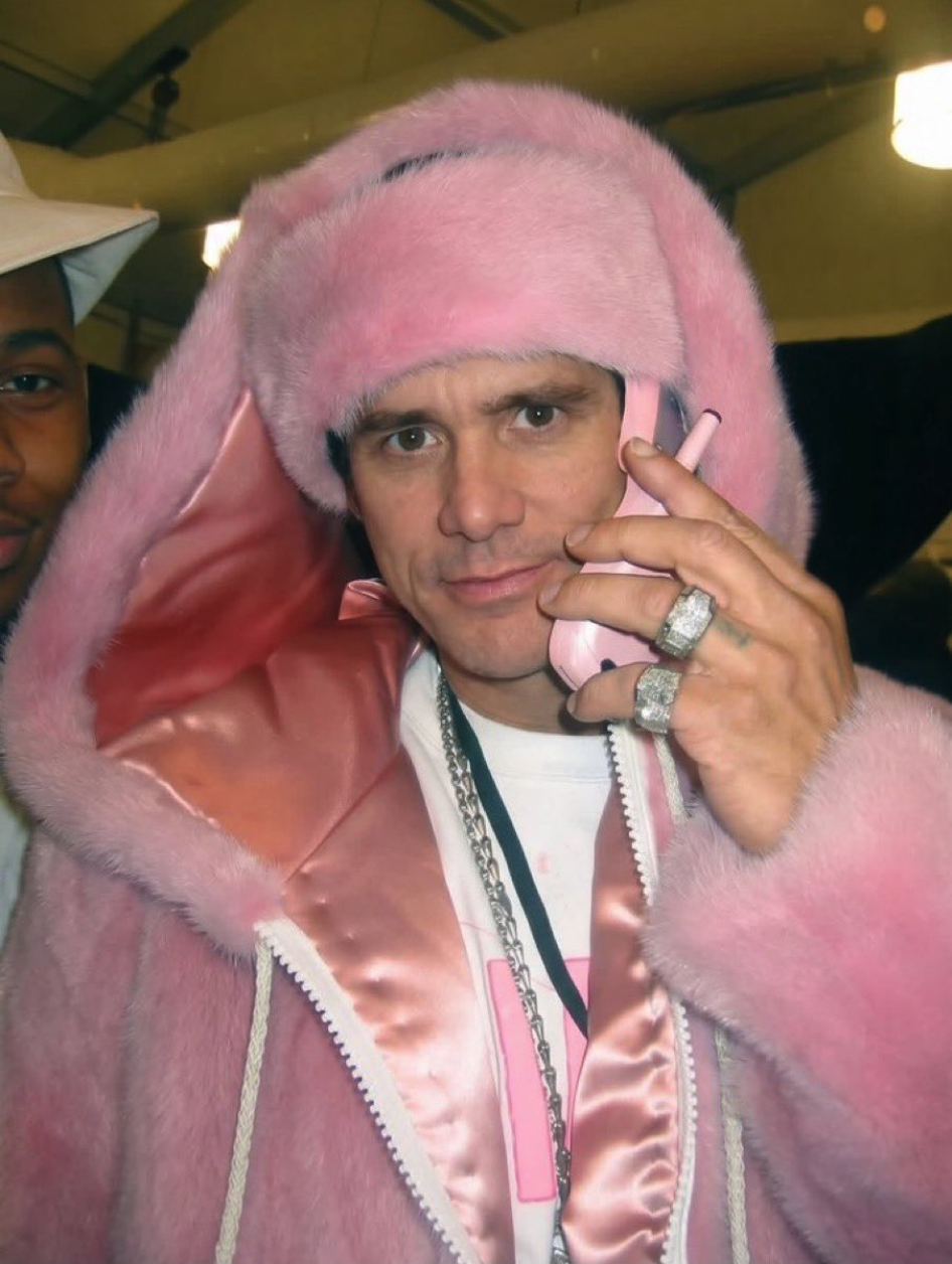 A man resembling Jim Carrey in a fluffy pink fur coat, holding a pink phone to his ear.