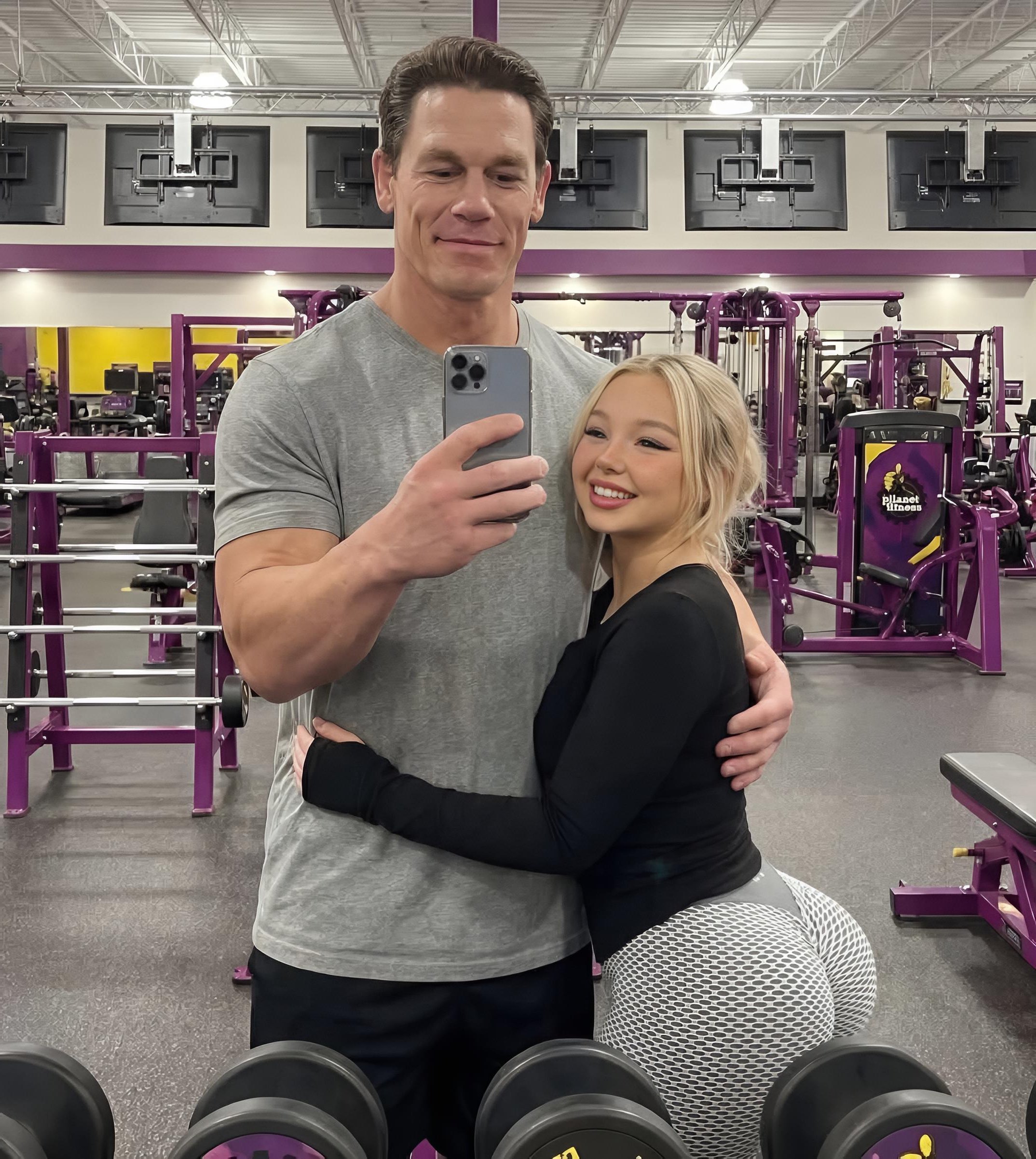 A tall man resembling John Cena taking a gym selfie while embracing a smiling blonde woman, with dumbbells in the foreground.