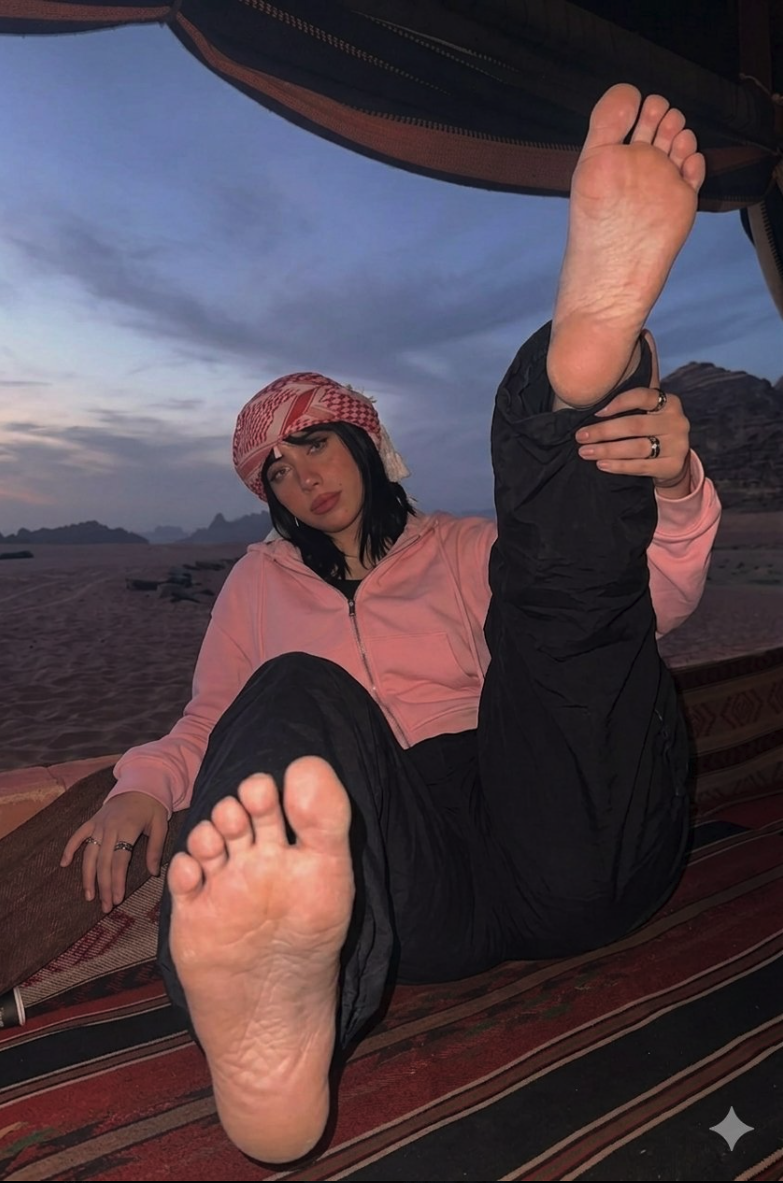 A woman resembling Kali Uchis reclining on a rug in a desert at dusk, wearing a red and white keffiyeh and pink hoodie.