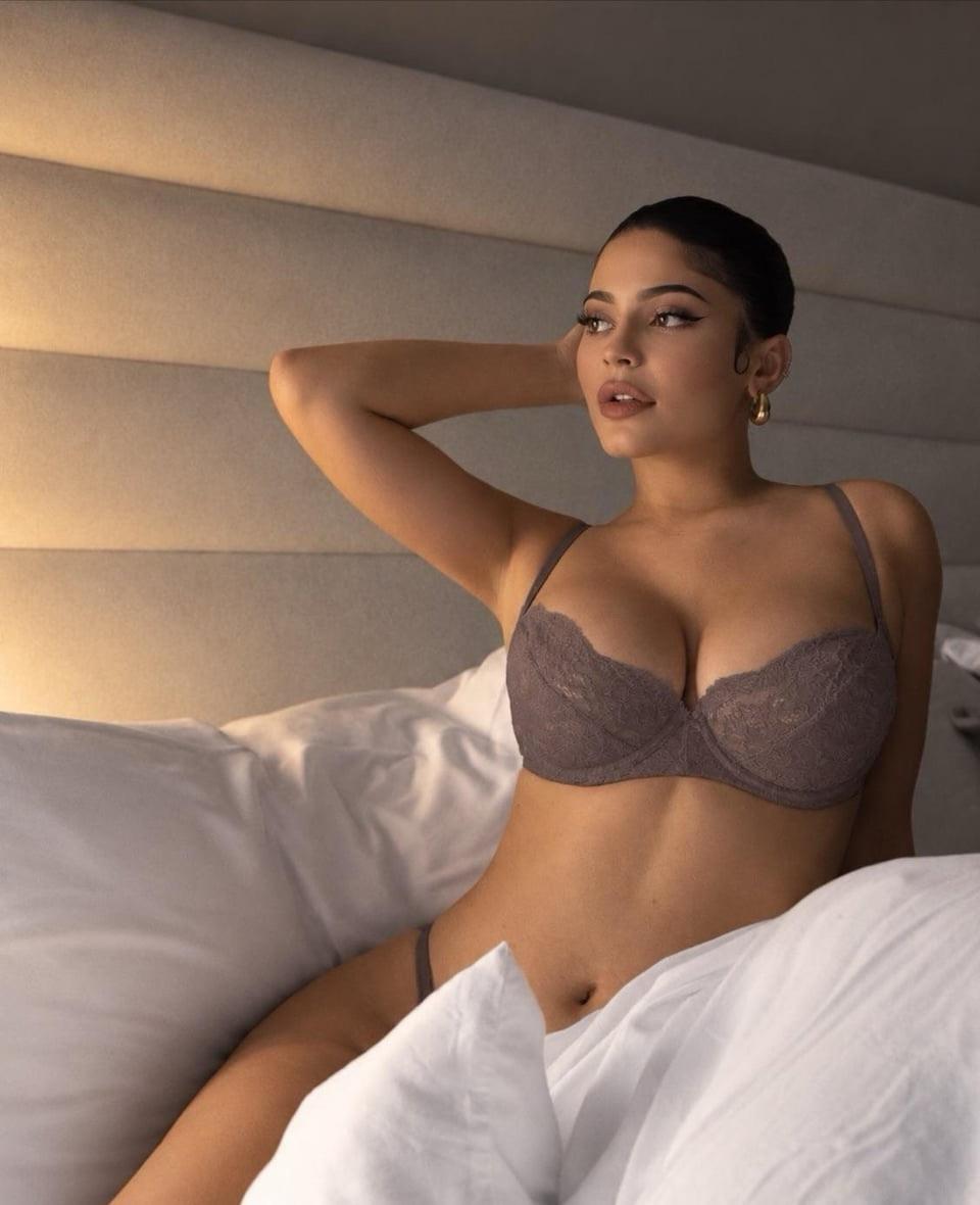 A woman resembling Kylie Jenner reclining on a bed in gray lace lingerie.