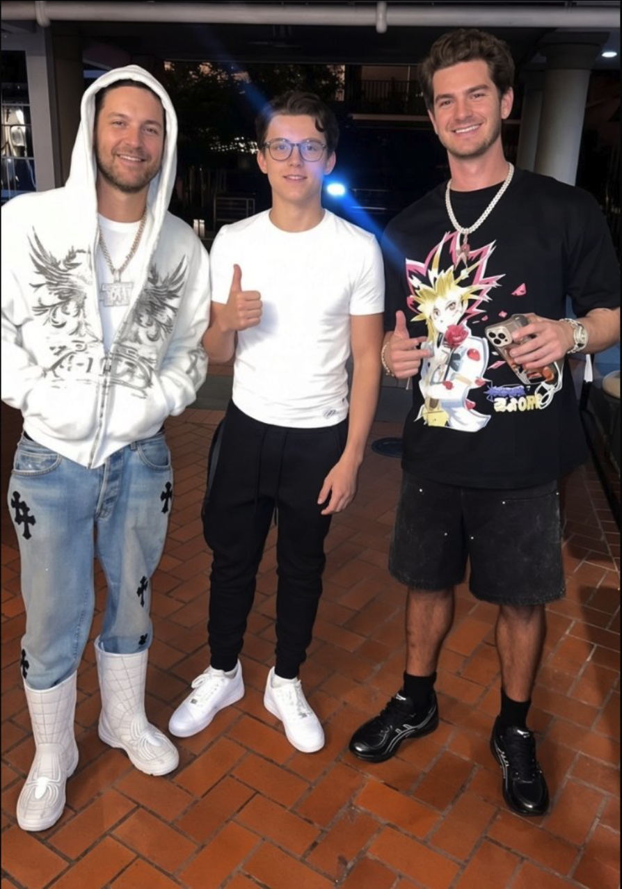 Three men resembling Tobey Maguire, Andrew Garfield, and Tom Holland posing together on a brick walkway at night.