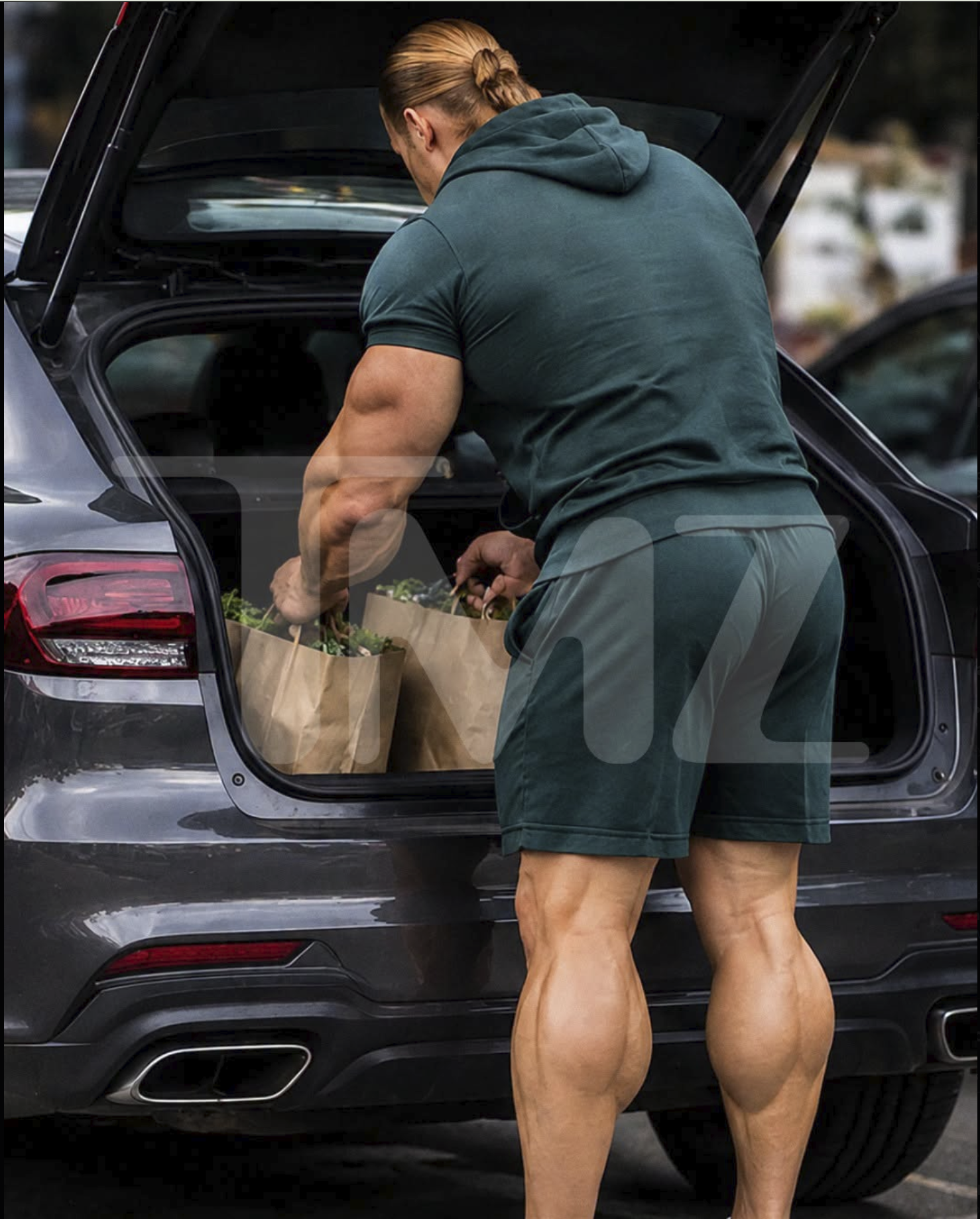A muscular man with a blond ponytail wearing athletic shorts and a t-shirt loading grocery bags into the open trunk of a dark SUV.