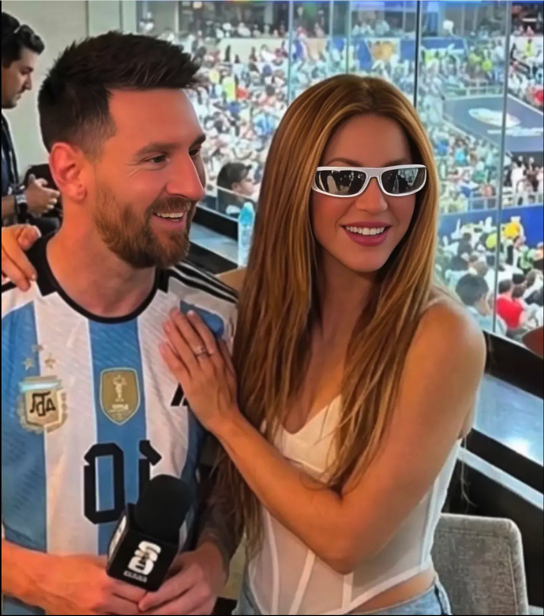 A man resembling Lionel Messi in an Argentina jersey and a woman resembling Shakira posing together in a stadium box.