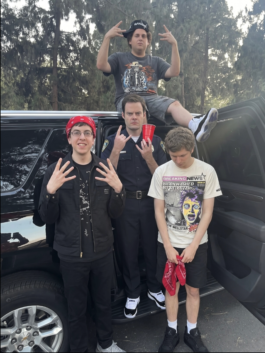 Four men resembling Christopher Mintz-Plasse, Bill Hader, Michael Cera, and Jonah Hill posing around a black SUV, with one wearing a police uniform.