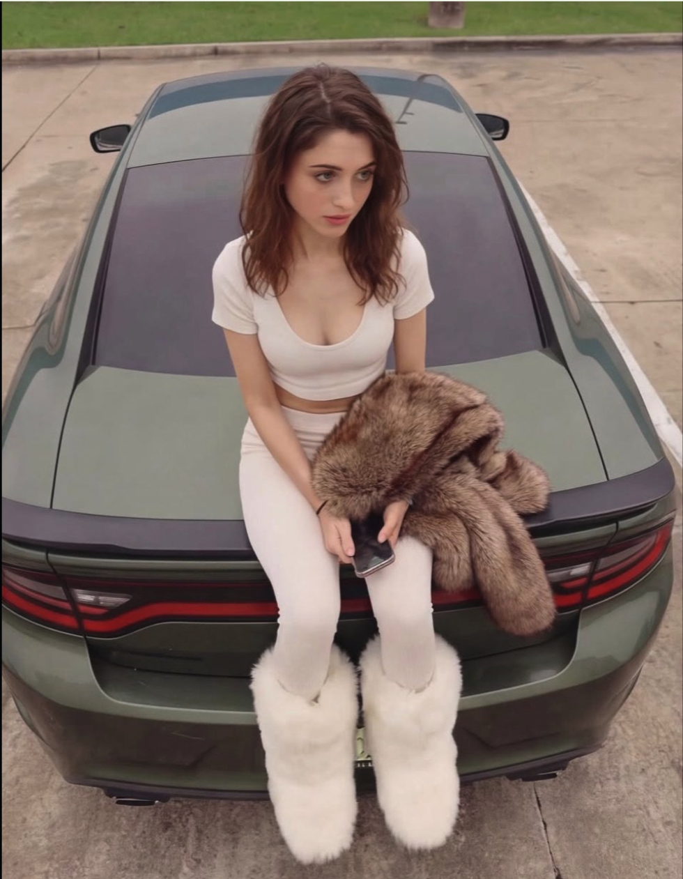 A woman resembling Natalia Dyer seated on the trunk of a dark green car, wearing a white crop top and white leggings.