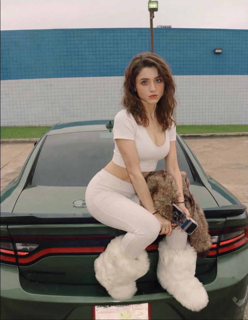 A woman resembling Natalia Dyer sitting on the trunk of a green Dodge Charger in a parking lot.