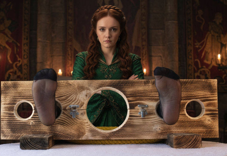 A woman resembling Olivia Cooke in a green medieval dress behind a wooden stocks device.