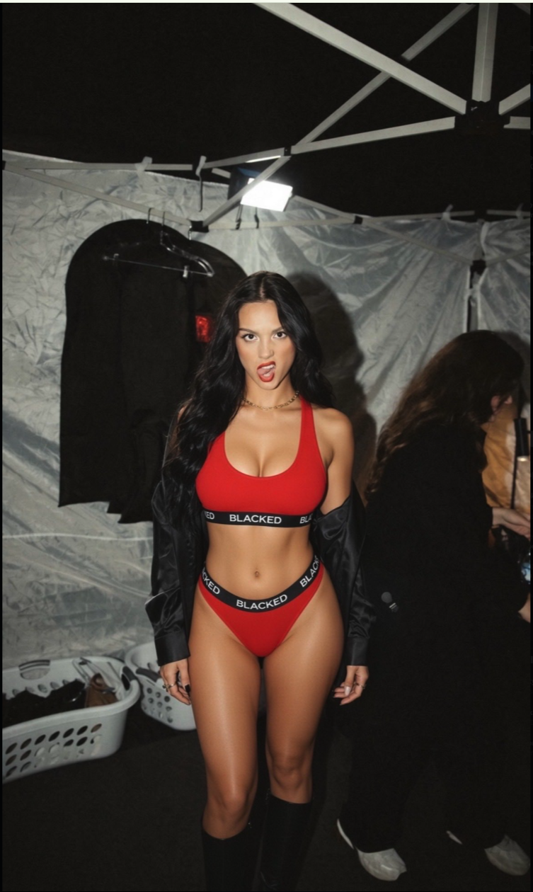 A woman resembling Paulina Krupińska wearing red branded underwear and a black jacket in a backstage tent.