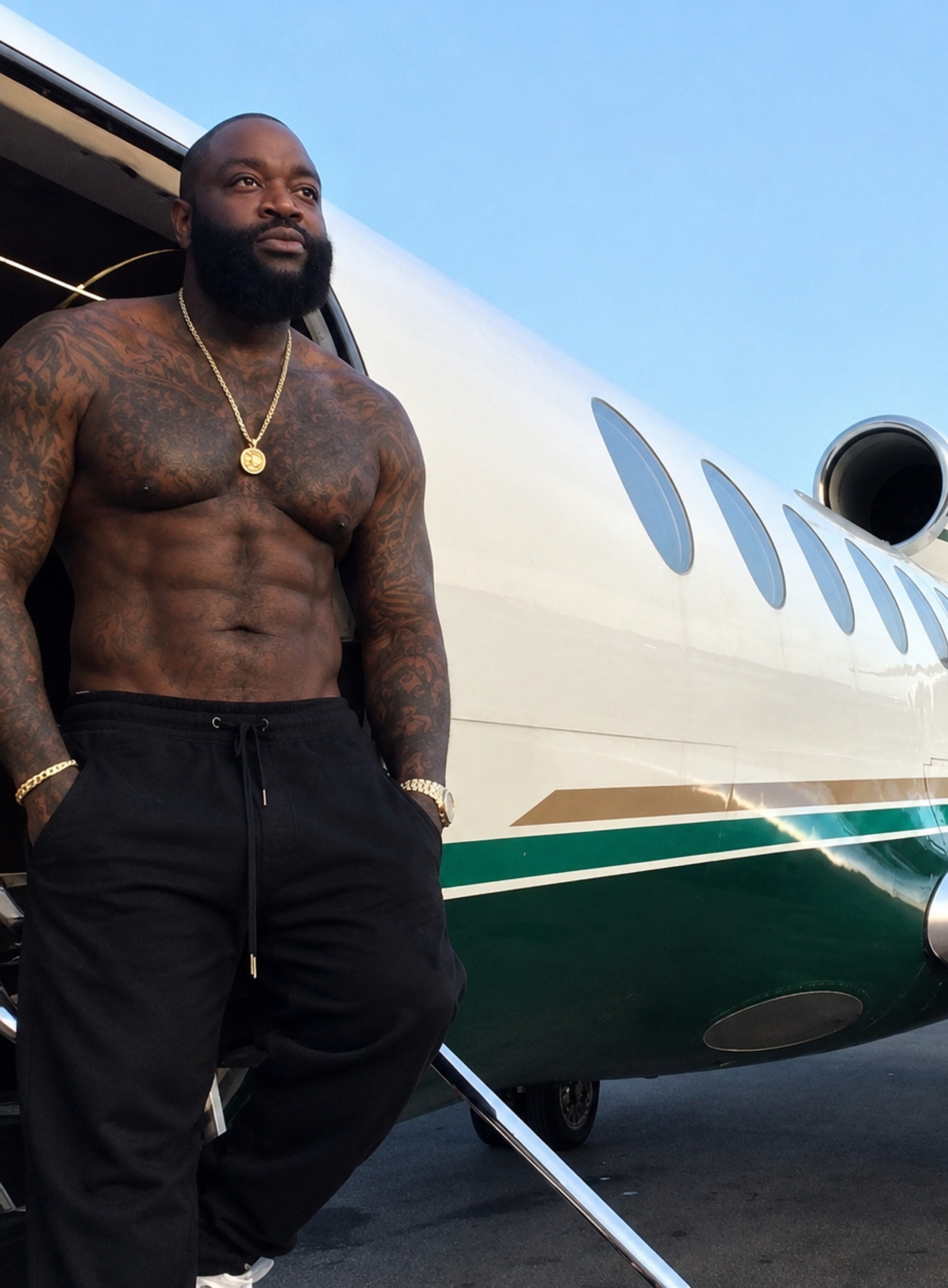 A shirtless man resembling Rick Ross standing beside a private jet on a tarmac.