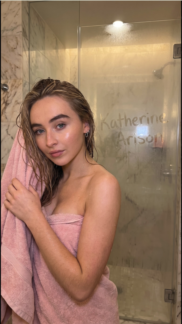 A young woman resembling Sabrina Carpenter wrapped in a pink towel inside a marble-tiled shower, with 'Katherine Arison' written on the glass.