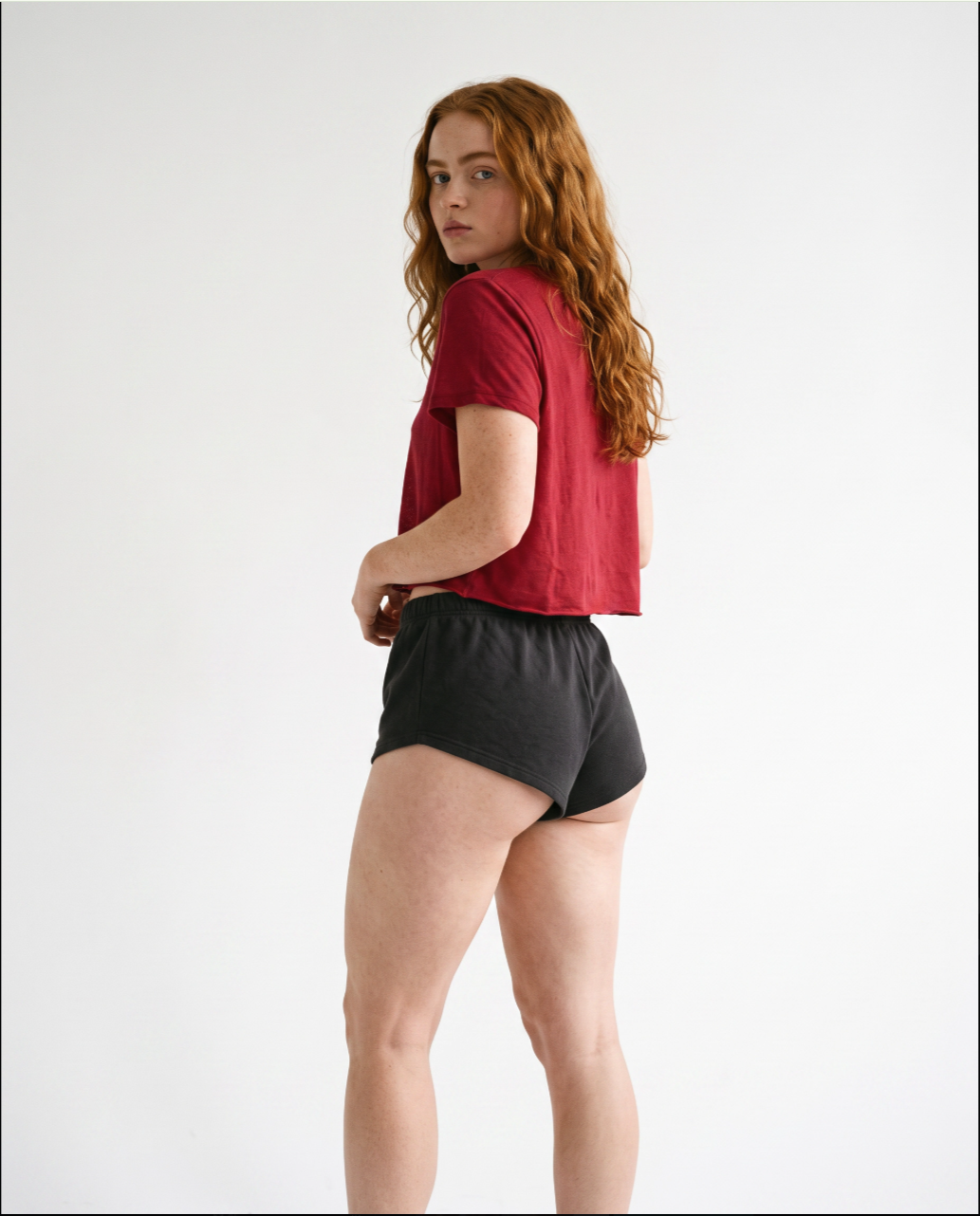 Sadie Sink a red t-shirt and black athletic shorts, looking over her shoulder against a white background.
