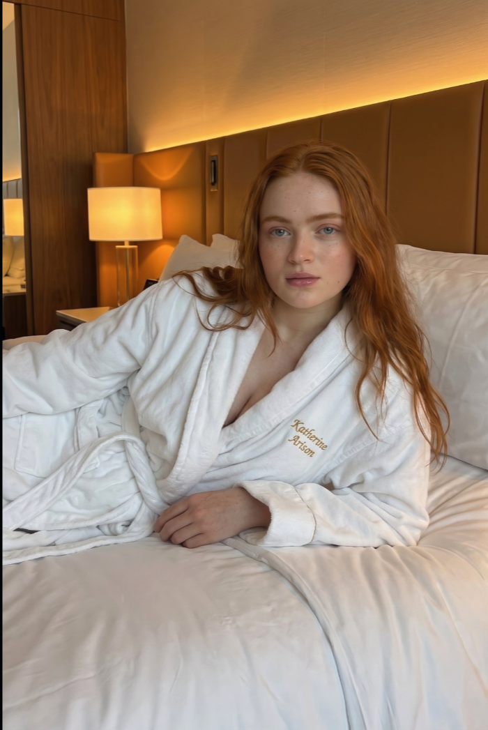 A woman resembling Sadie Sink lying on a hotel bed in a white bathrobe embroidered 'Katherine Arison'.