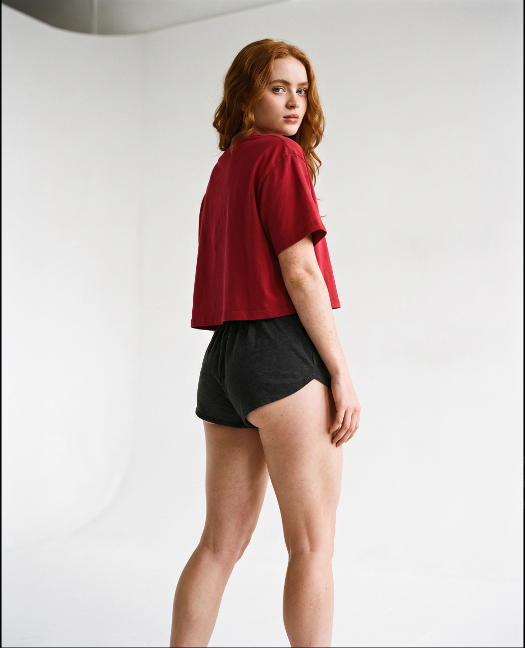 A woman resembling Sadie Sink posing in a red top and dark shorts against a light backdrop.