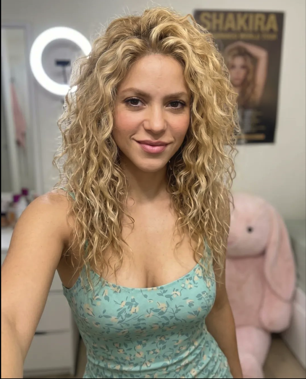 A woman resembling Shakira posing indoors in a teal floral dress, with a Shakira tour poster on the wall behind her.