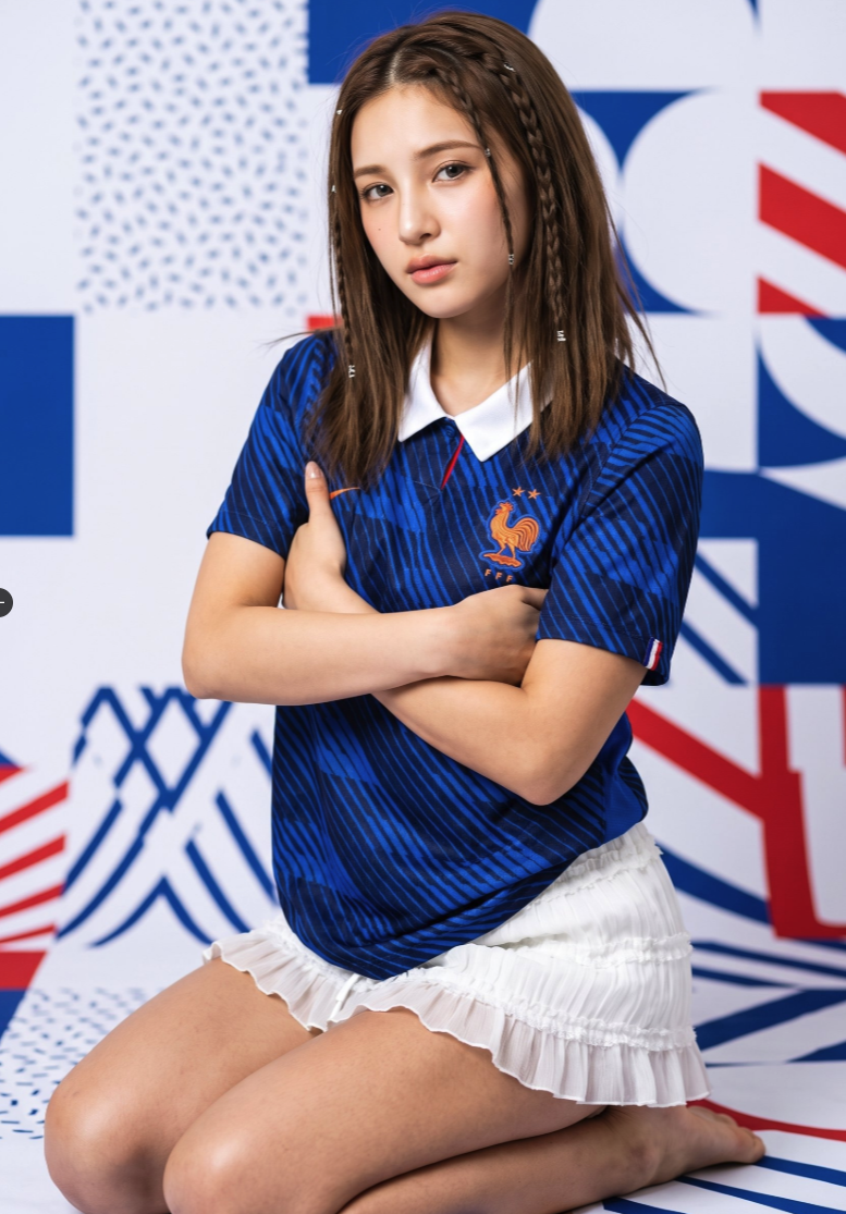 A woman wearing a dark blue France national football jersey and a white pleated skirt, seated in front of a red, white, and blue patterned background.