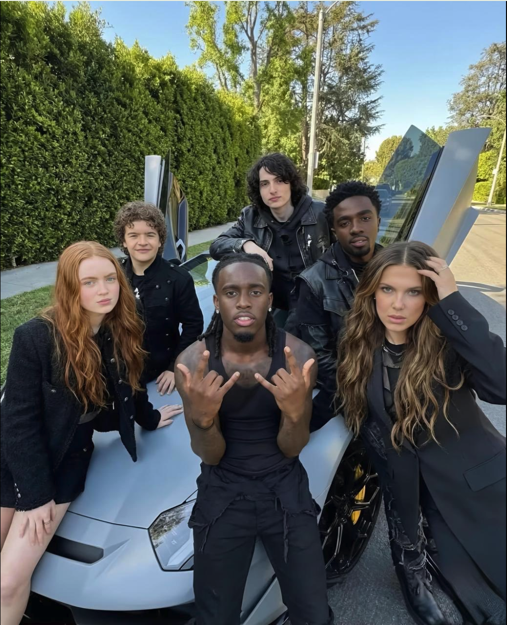 Six people resembling Stranger Things cast members gathered around a light blue Lamborghini with scissor doors open outdoors.